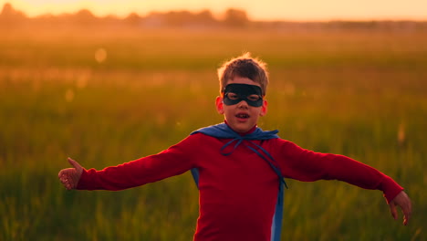 A-boy-in-a-suit-and-a-superhero-mask-runs-across-the-field-at-sunset-on-the-grass-dreaming-and-imagining-himself-a-hero-and-a-winner.-Fun-and-happy-family